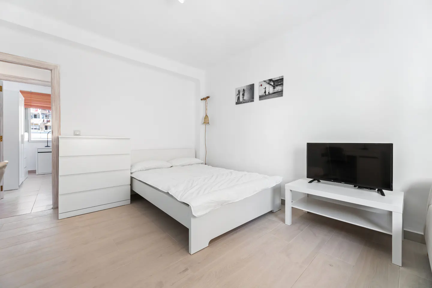 Bright, modern bedroom with white walls, light wood floors, and a white bed. A TV sits on a white stand, and a dresser is next to the bed.