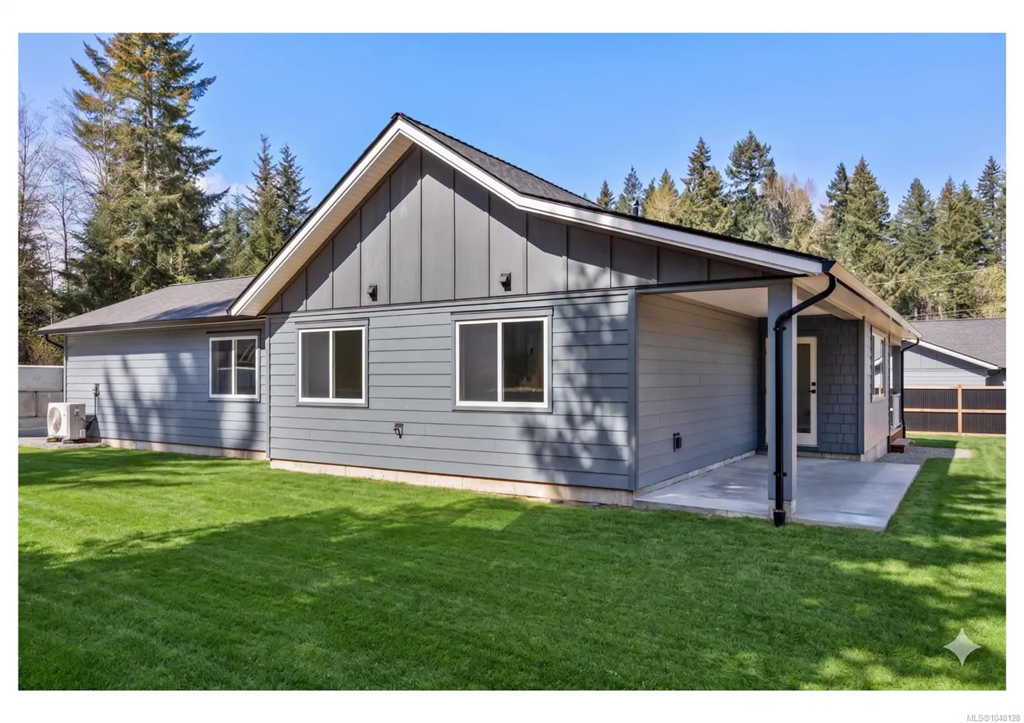 Exterior view of a modern gray house with a covered patio, large green lawn, and a backdrop of tall evergreen trees.