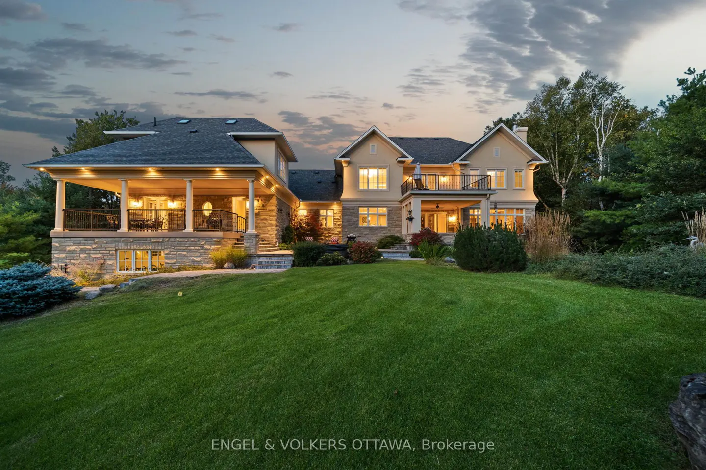 A large, two-story tan house with a stone base and a green lawn at dusk.