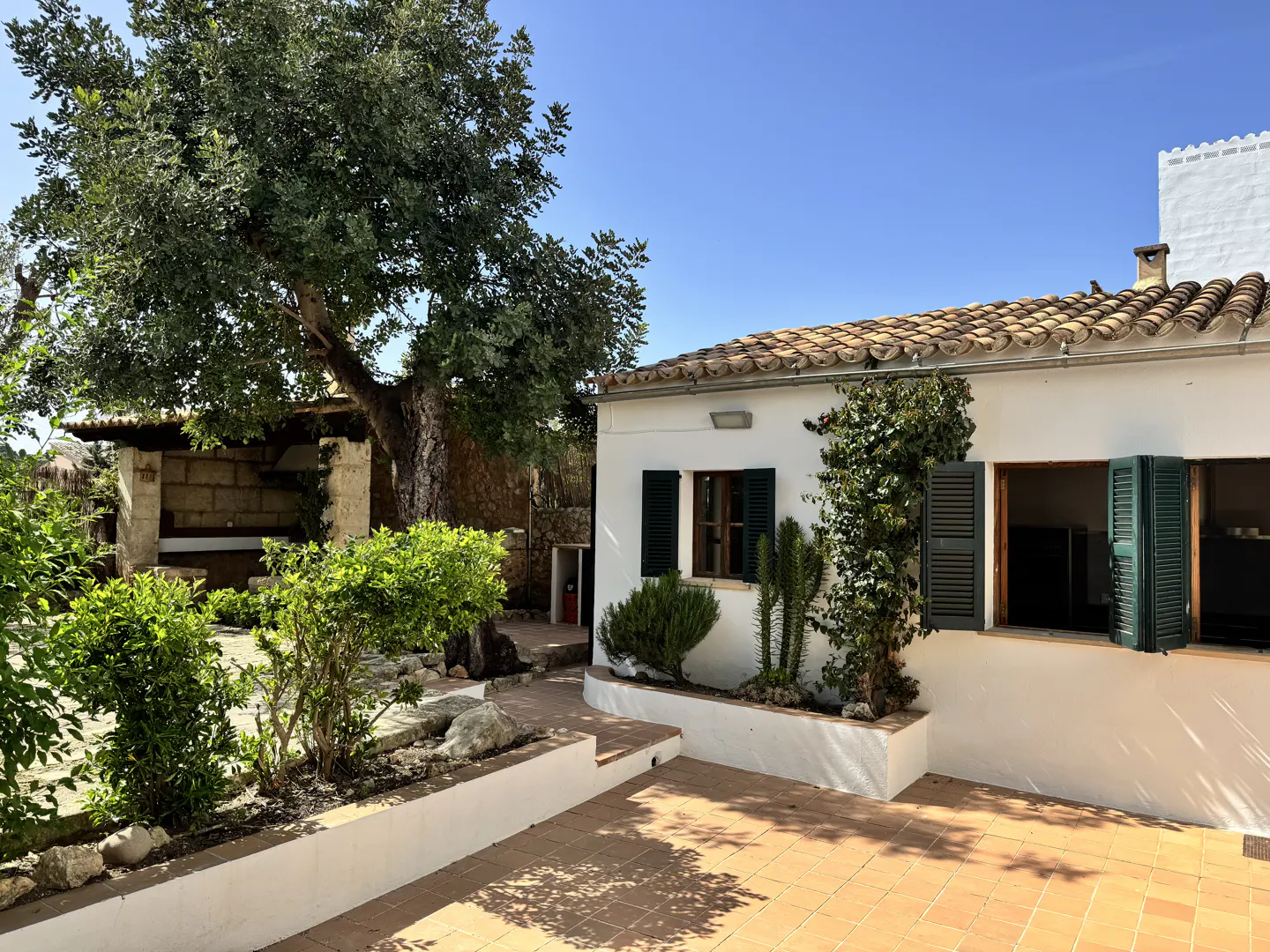 Exterior view of a white building with green shutters, a terracotta tile roof, and a brick patio. Lush greenery surrounds the building.
