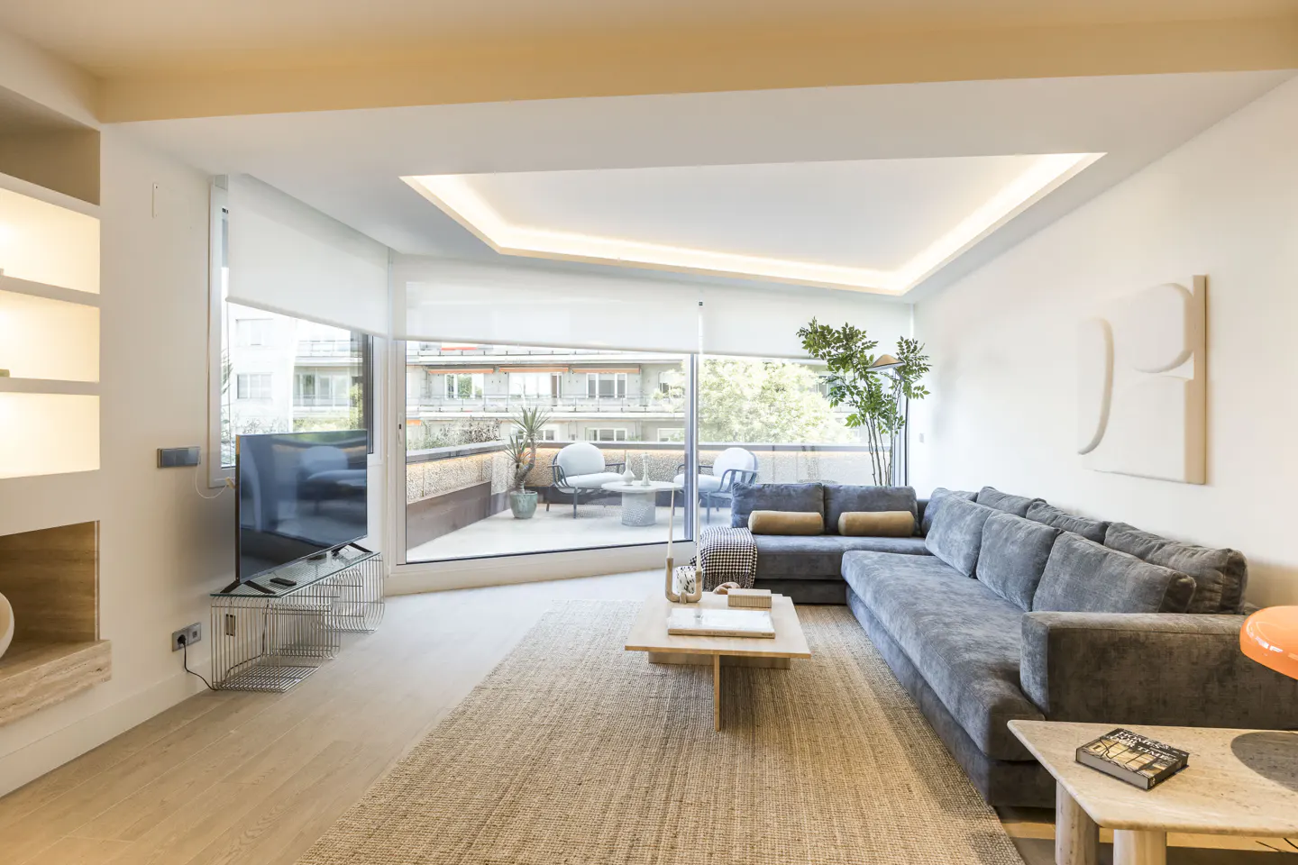 Modern living room with a large grey sectional sofa, wooden coffee table, and floor-to-ceiling windows leading to a balcony.
