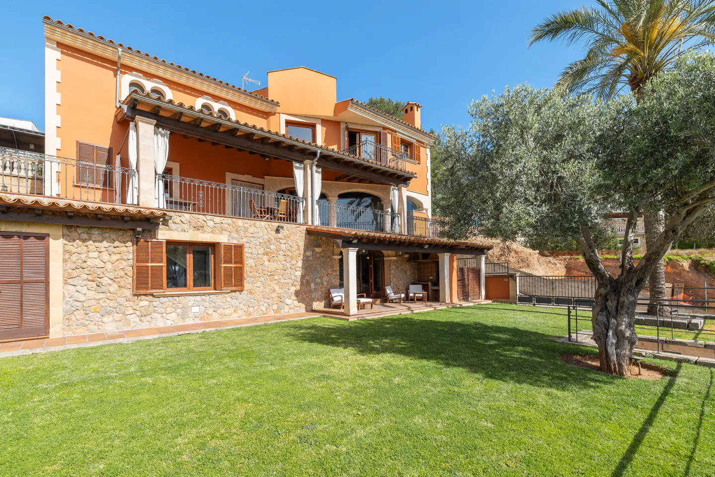 Exterior view of a two-story orange house with stone accents, balconies, a green lawn, and trees under a clear blue sky.