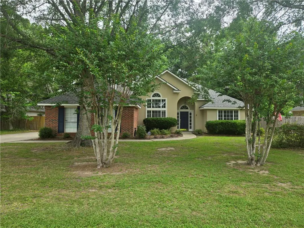 A tan single-story house with a brick accent wall and a dark gray roof is surrounded by green trees and a lawn.