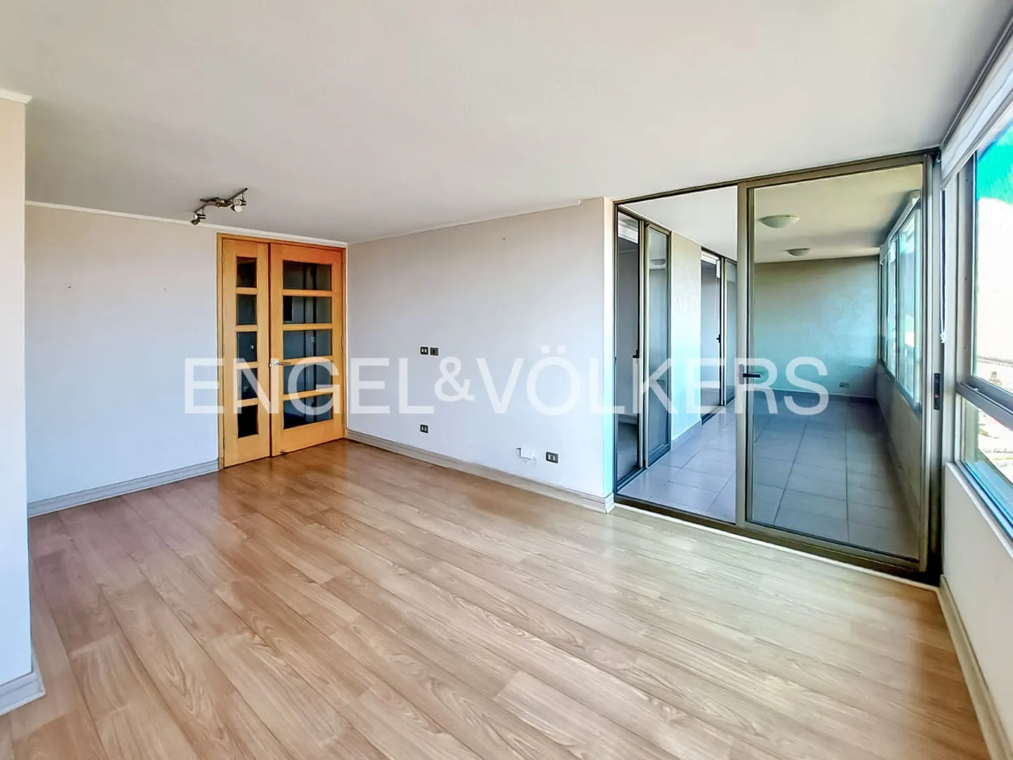 Empty room with wood floors, white walls, and glass doors to a sunroom. Engel & Völkers logo on the wall.