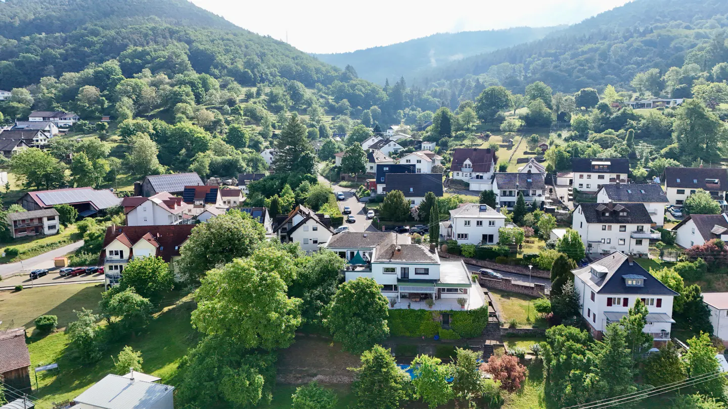 Aerial view of a hillside town with houses nestled among green trees and mountains in the background.