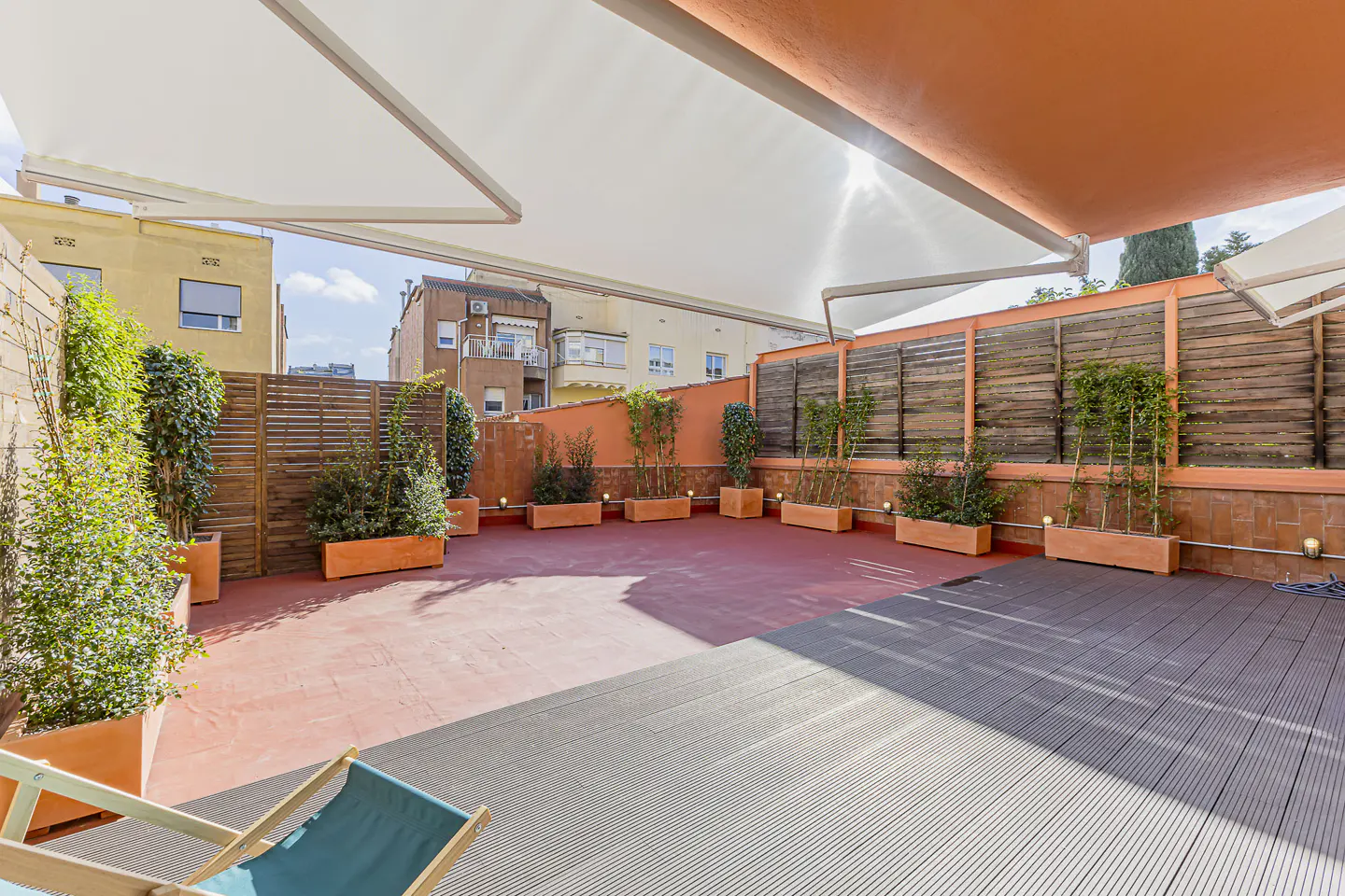Outdoor patio with gray composite decking, red painted concrete, and orange planters with greenery. White awnings provide shade.