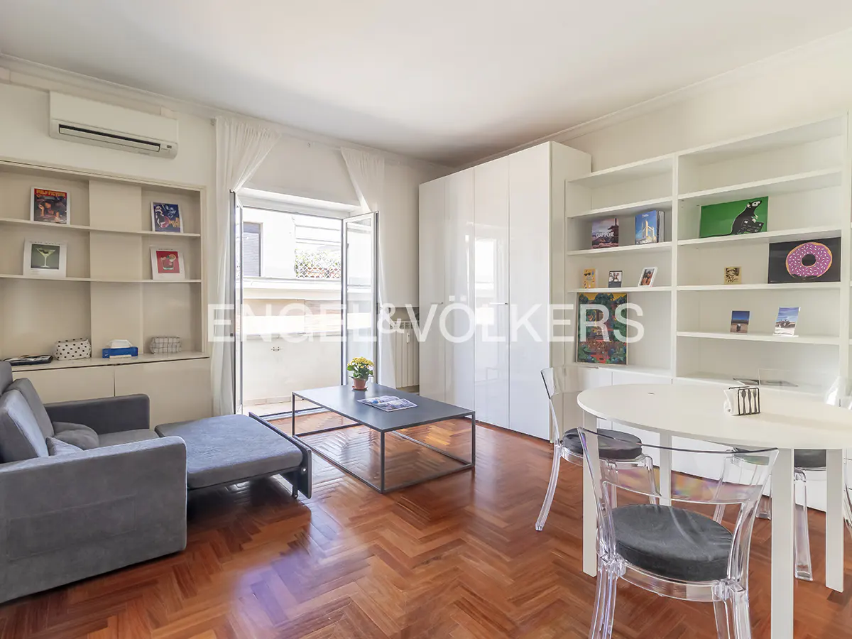 Bright living room with herringbone wood floors, grey sofa, black coffee table, and white shelving units. Balcony doors open to the outside.