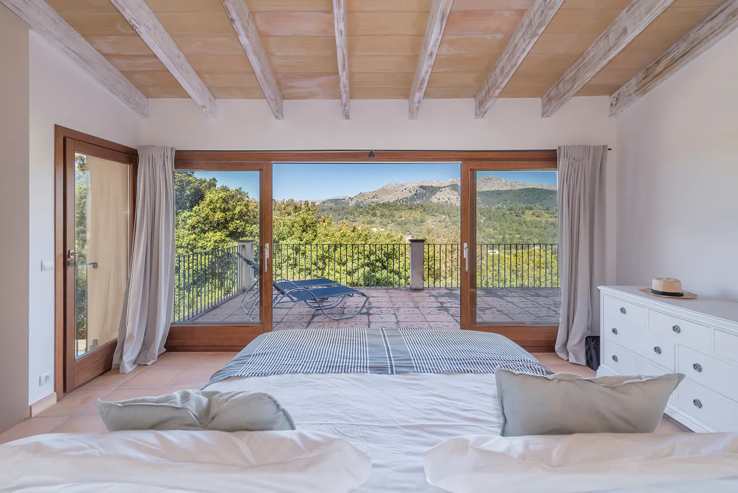 Bedroom with white linens, light blue pillows, and a blue striped blanket. A terrace with a lounge chair overlooks a green landscape.