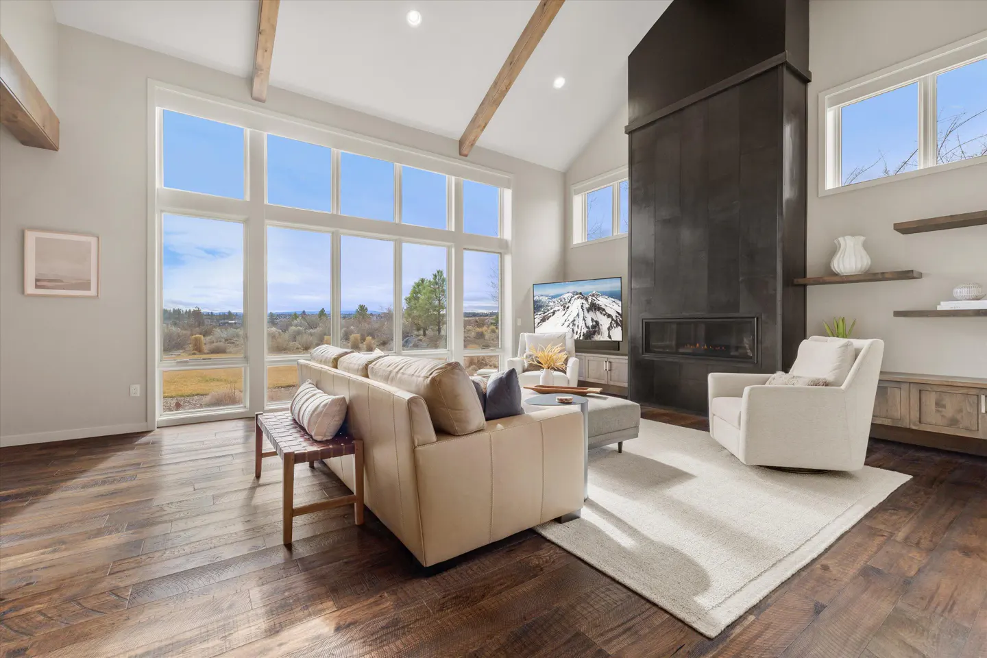 Living room with hardwood floors, large windows, and a stone fireplace. Beige sofa and chair sit on a light rug. Beamed ceiling.