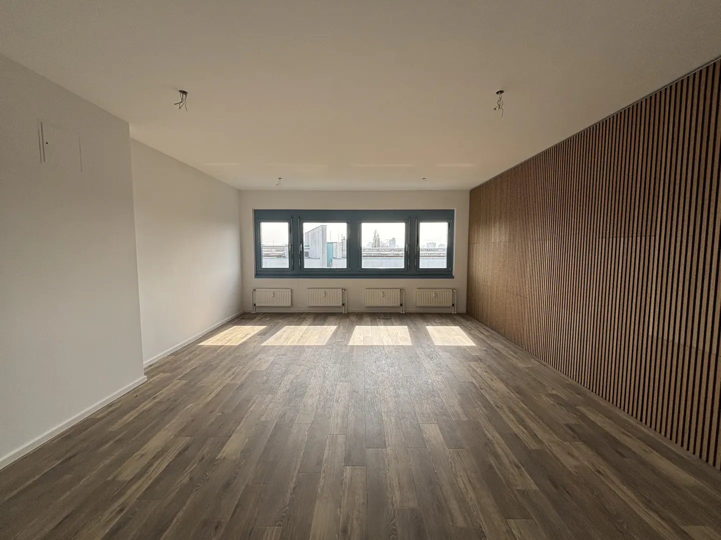 Empty room with wood floors, white walls, and a wood slat wall. Windows overlook a city skyline and water.