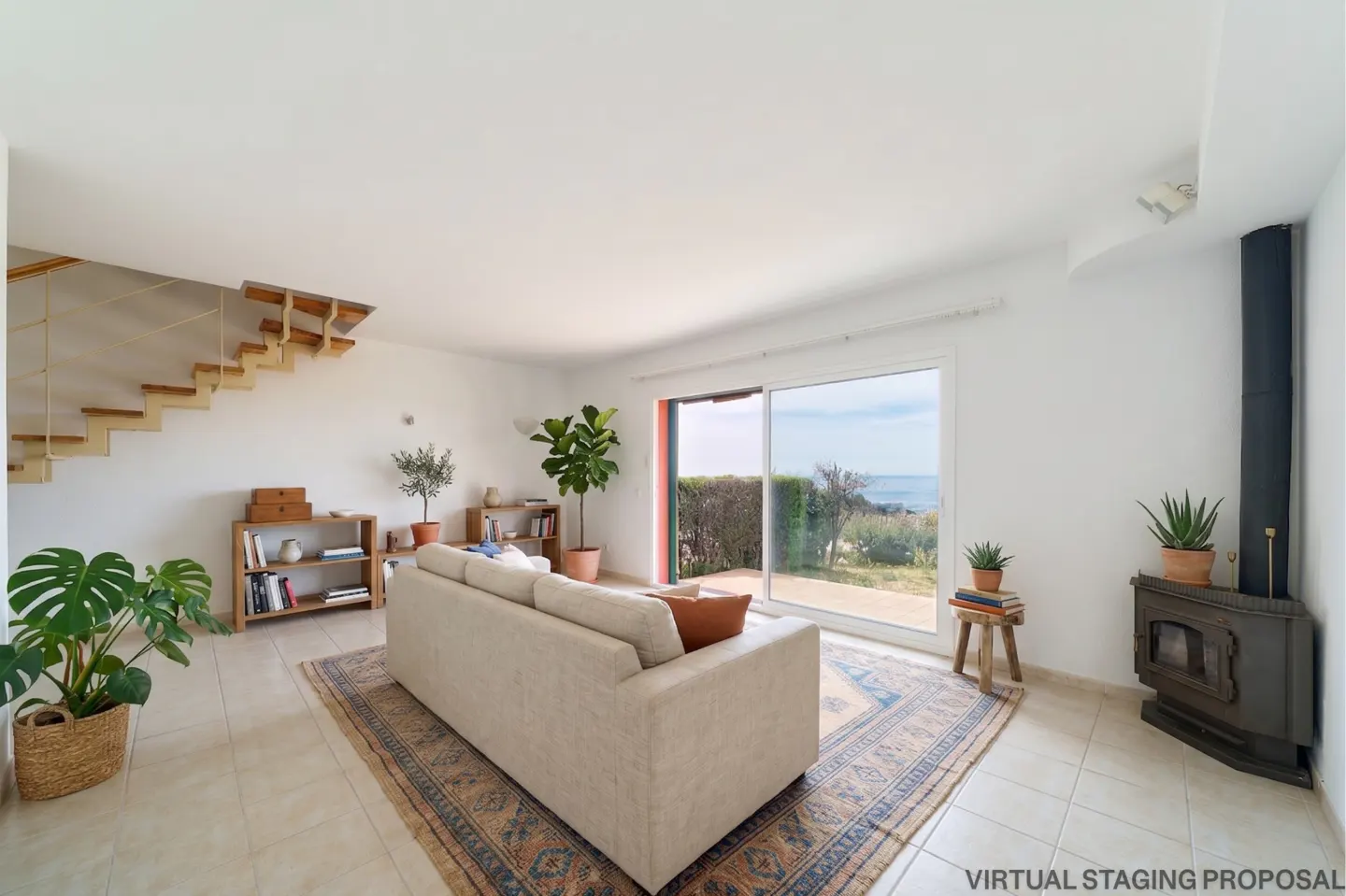 Bright living room with a beige sofa on a patterned rug, plants, and a view of the ocean through a sliding glass door.