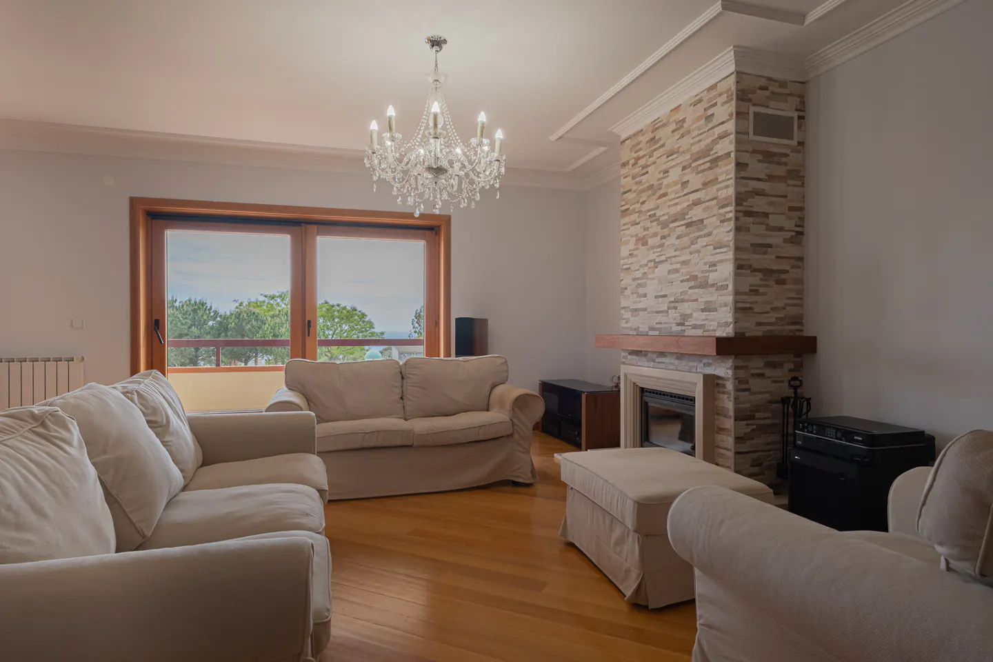 Living room with hardwood floors, white sofas, and a stone fireplace. A crystal chandelier hangs from the ceiling. Large windows offer a view of trees.