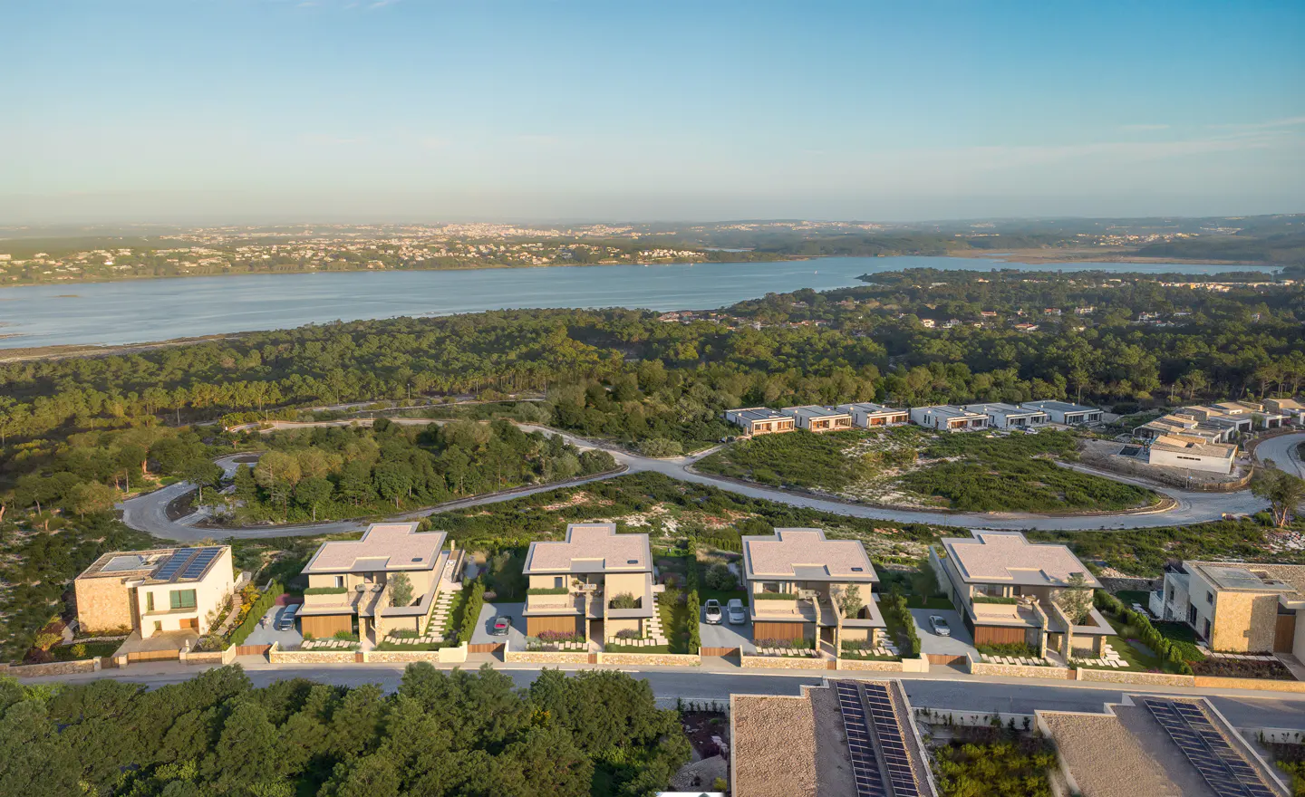 Aerial view of modern beige houses with flat roofs, surrounded by green trees and a blue lake in the background.