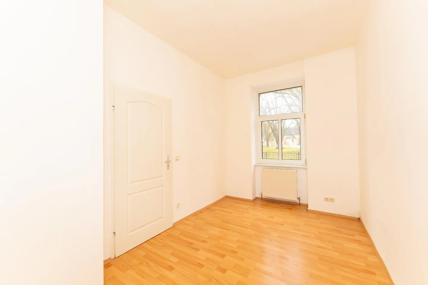 Bright, empty room with wood floors, white walls, a window showing trees, and a white door.