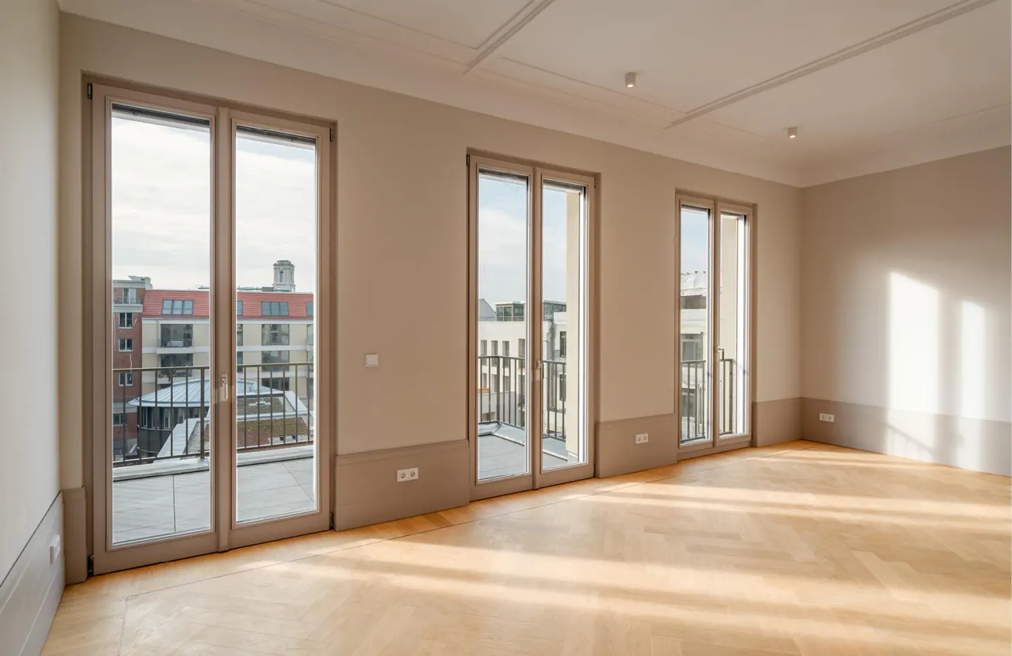 Bright, empty room with light wood herringbone floors, beige walls, and three glass balcony doors overlooking a city.