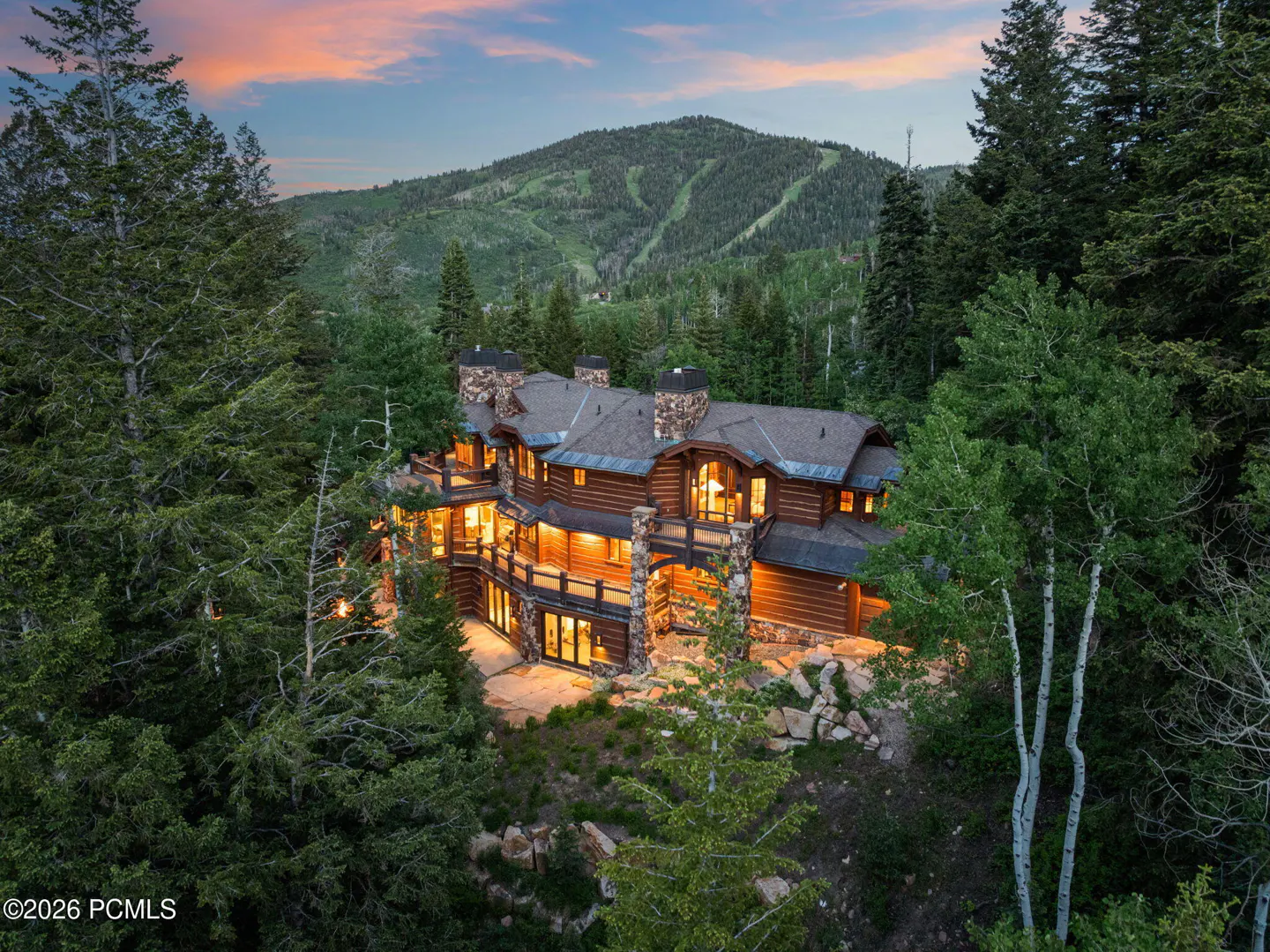 A large, illuminated log cabin with stone chimneys sits nestled in a forest at dusk. A mountain is visible in the background.