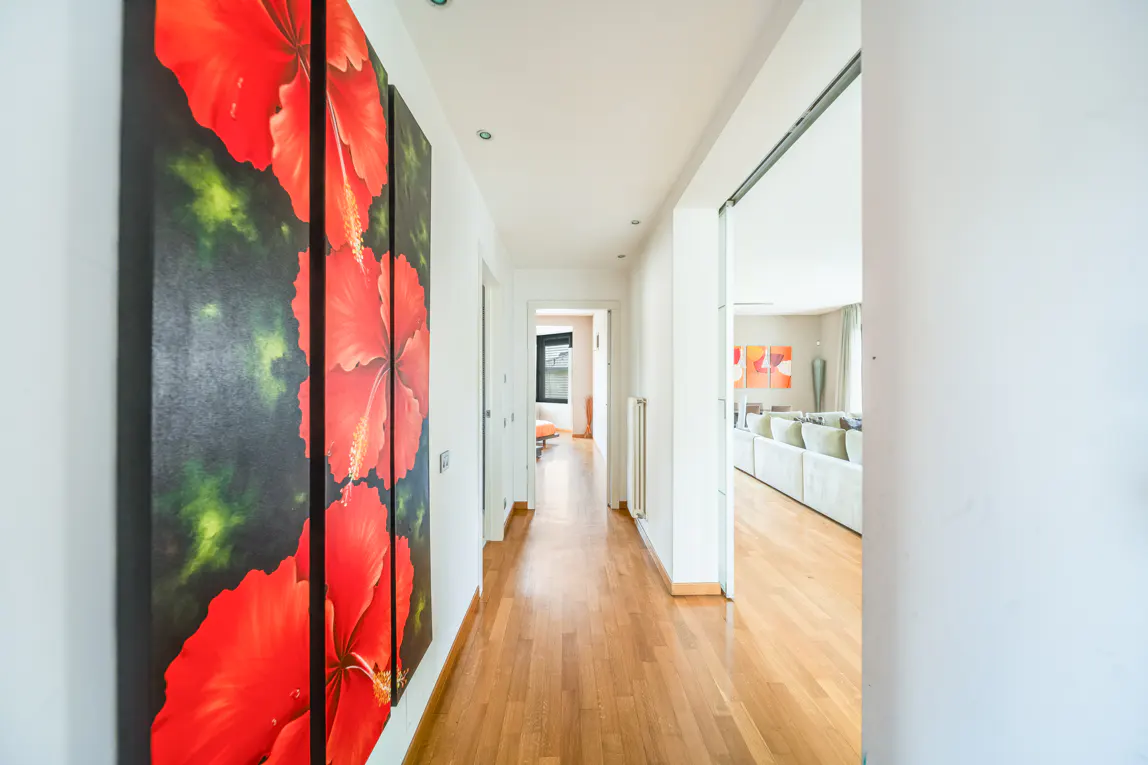 Hallway with hardwood floors and white walls. A large red flower triptych hangs on the left wall. A living room is visible through a doorway.