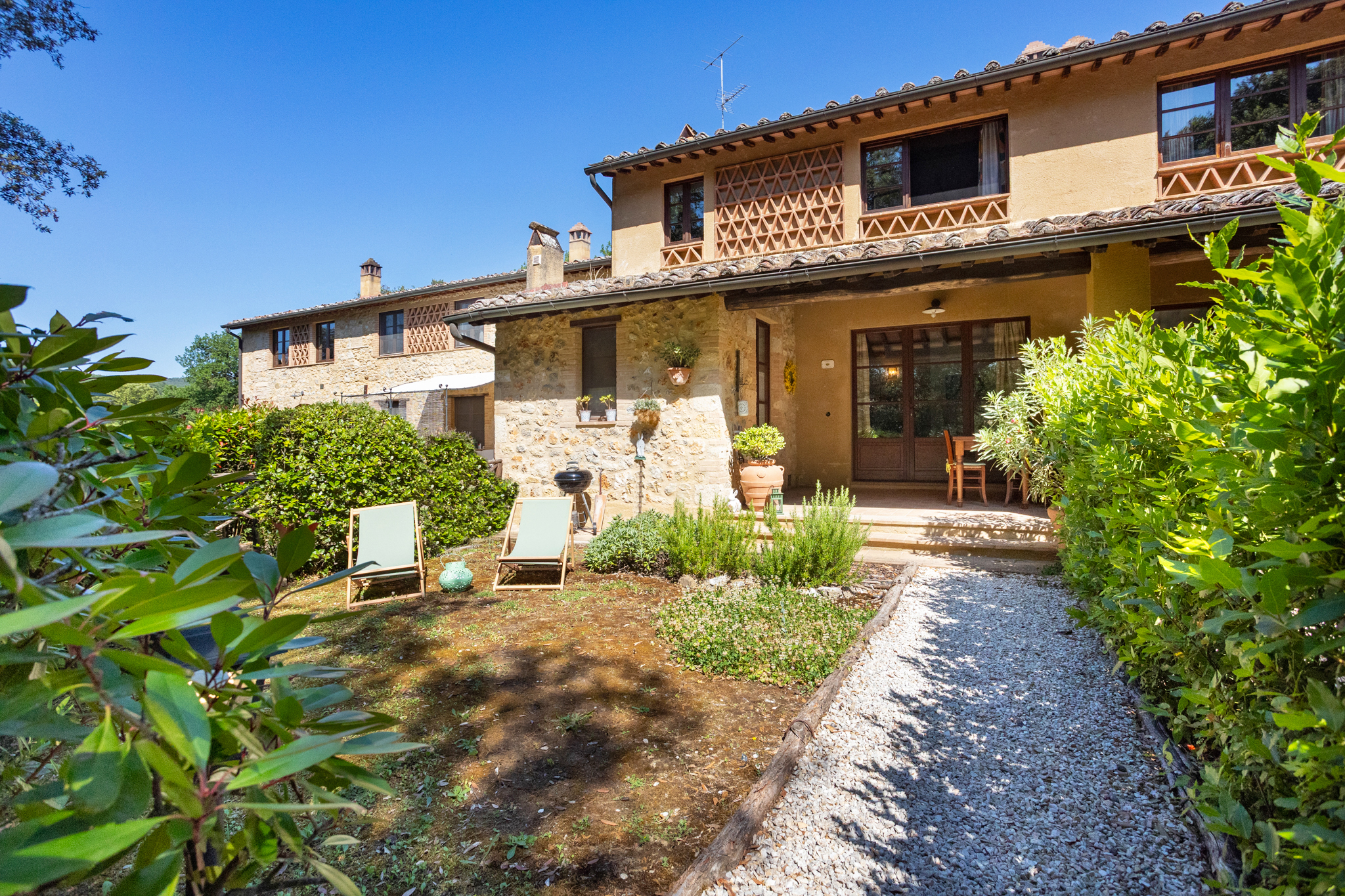 A rustic stone villa with a gravel path, green lawn, lounge chairs, and lush garden under a clear blue sky.