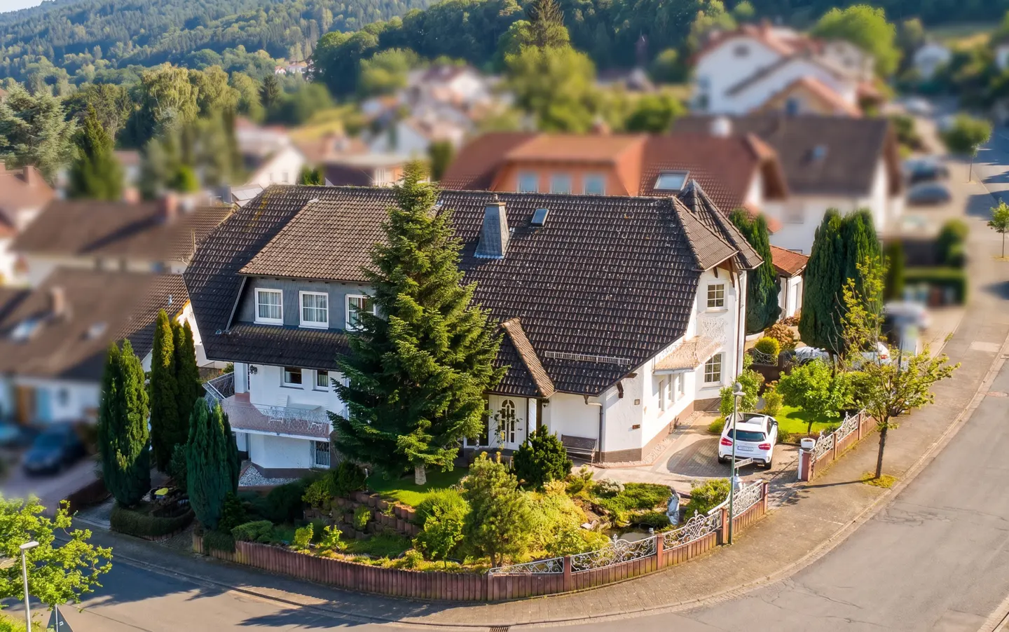 Aerial view of a white house with a brown roof, surrounded by trees and a garden, with a car parked nearby.