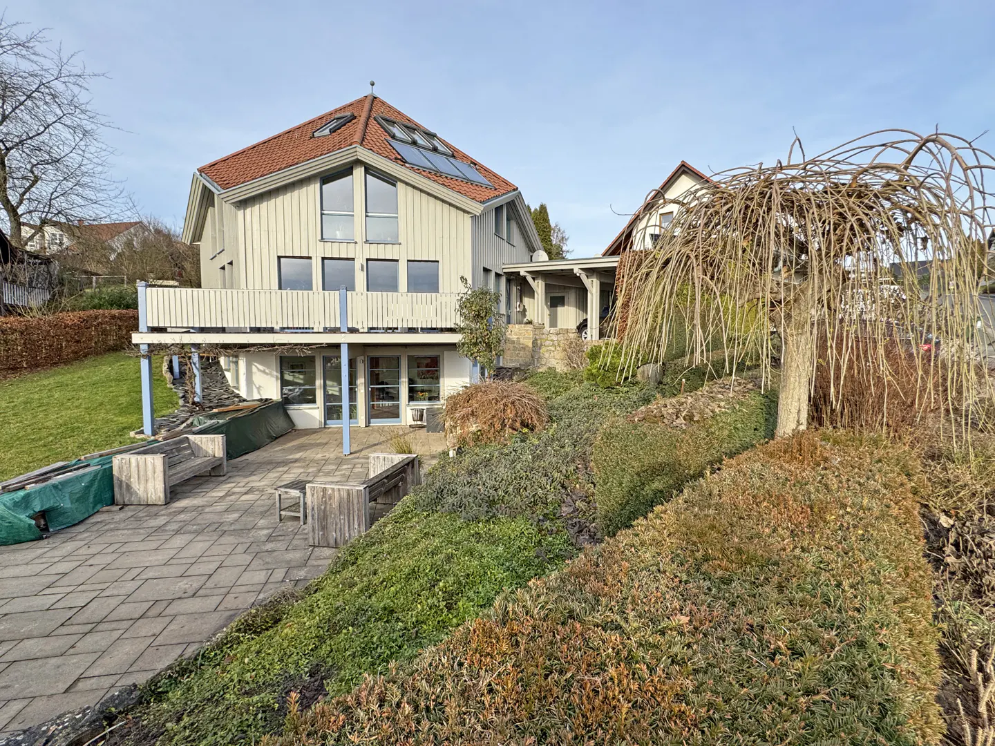 A two-story house with a red tile roof, light gray siding, and a blue-trimmed balcony overlooks a stone patio and landscaped hillside.