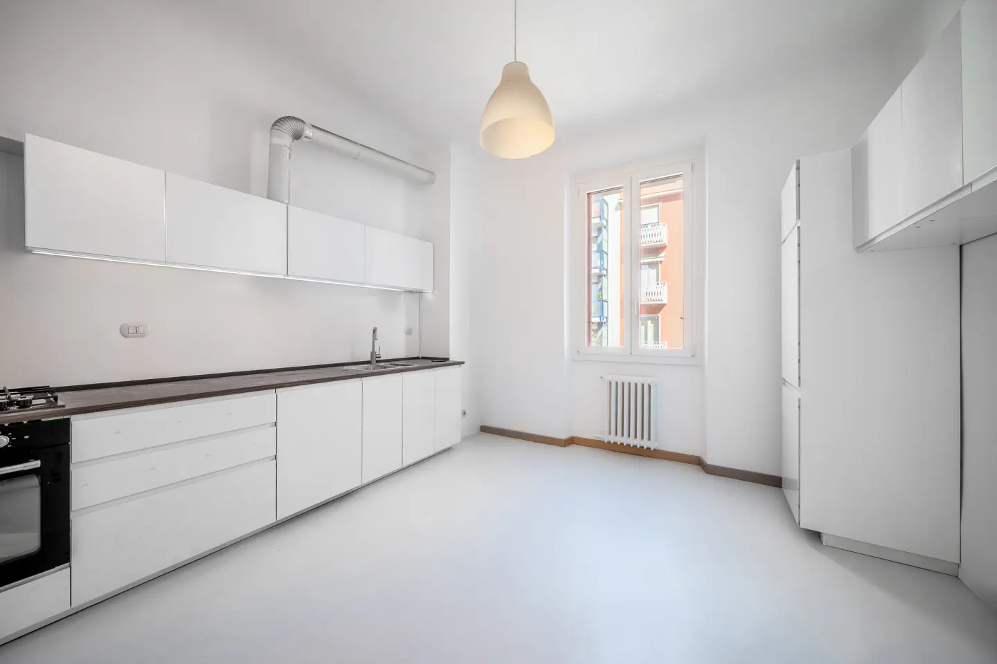 Bright, modern kitchen with white cabinets, dark countertops, and a window showing a building exterior. A pendant light hangs above.