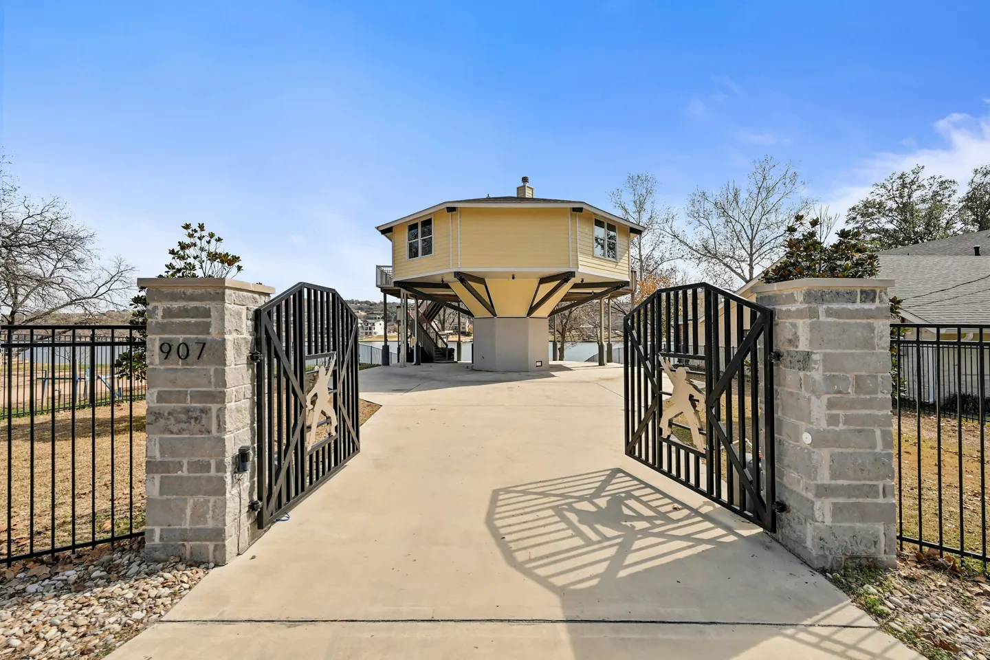 View of a yellow, octagonal house on stilts, seen through open black metal gates with stone pillars. The house overlooks a lake under a blue sky.