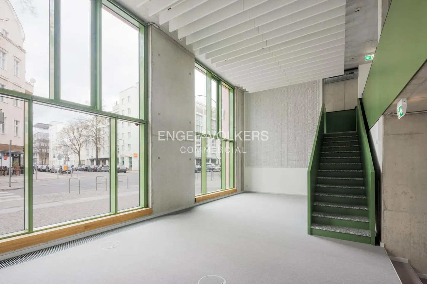 Empty commercial space with large windows, gray floor, concrete walls, and green-trimmed staircase. Street view visible through windows.