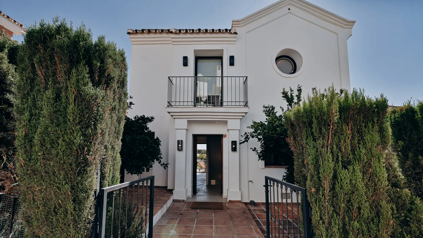 Two-story white house with a balcony and a round window, surrounded by green trees and a terracotta tile walkway.