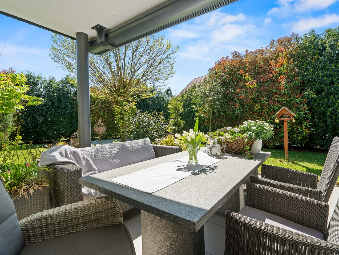 Outdoor patio with gray wicker furniture, a stone table with flowers, and a lush green garden backdrop under a bright blue sky.