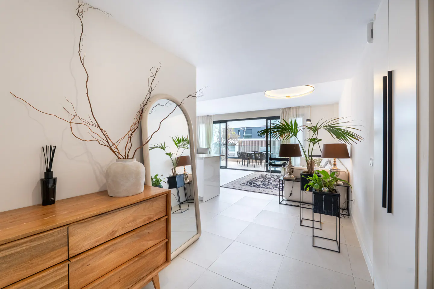 Bright apartment entryway with white tile floors, a wooden dresser, and a large arched mirror reflecting the living room and balcony.