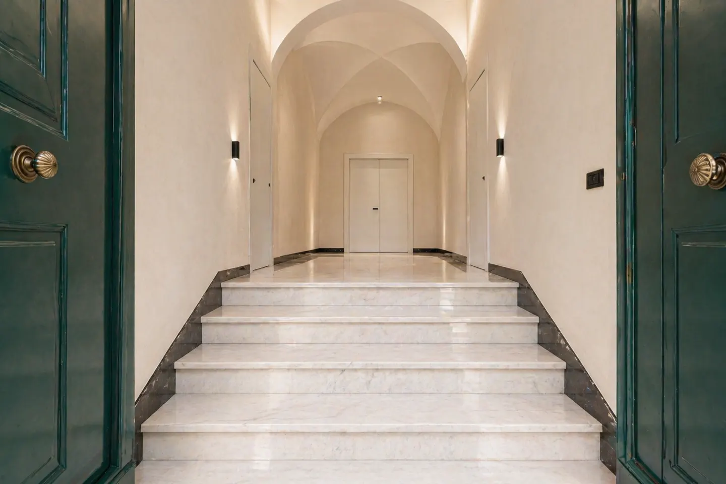 A marble staircase leads up to a vaulted hallway with white walls, modern sconces, and dark green doors on either side.
