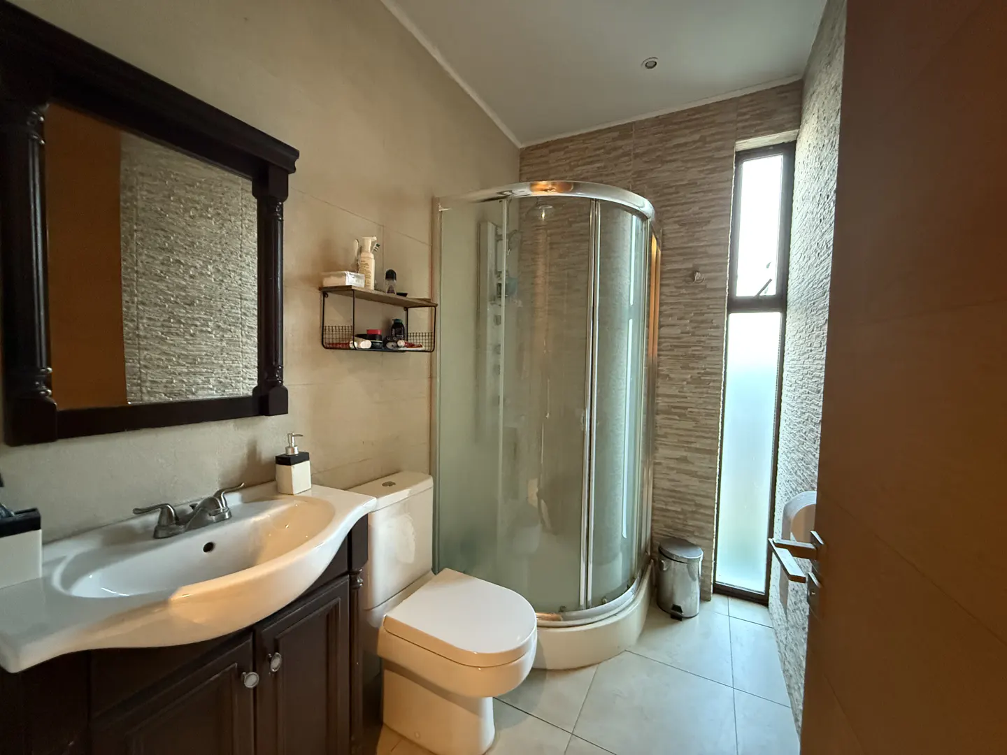 Bathroom with a dark wood vanity, white sink, toilet, and glass shower. A mirror hangs above the sink. A window is to the right of the shower.