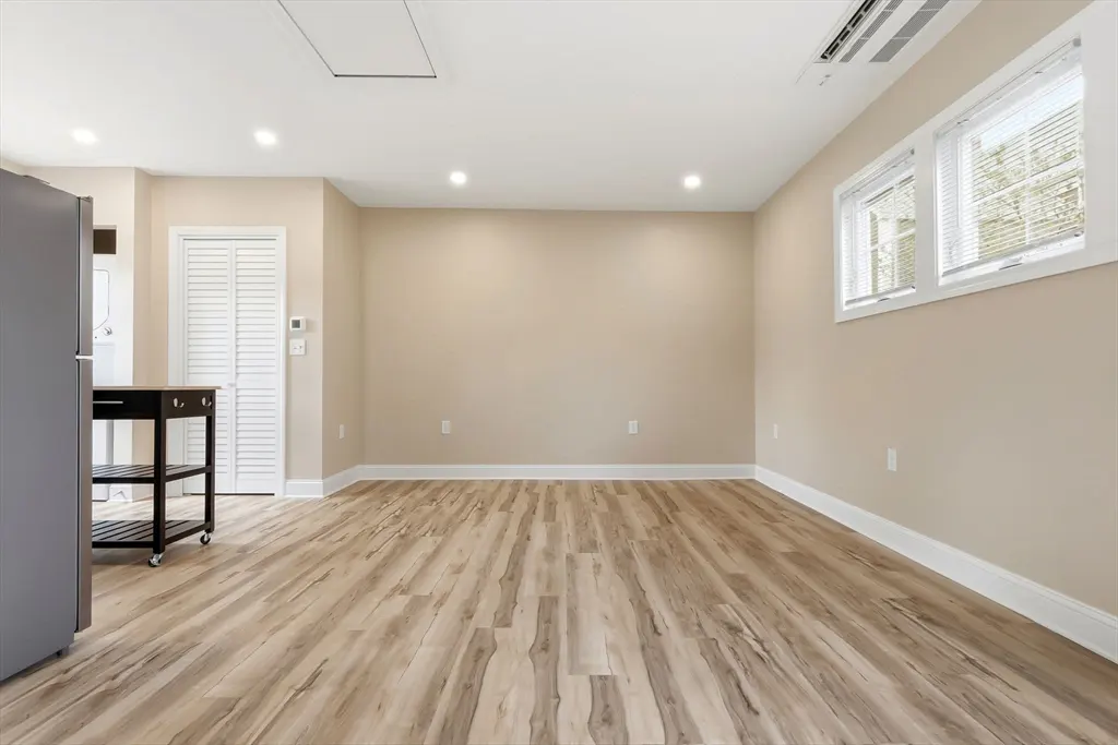 Bright, empty room with wood-look floors, beige walls, and white trim. A gray refrigerator and black rolling cart are on the left. Two windows are on the right.