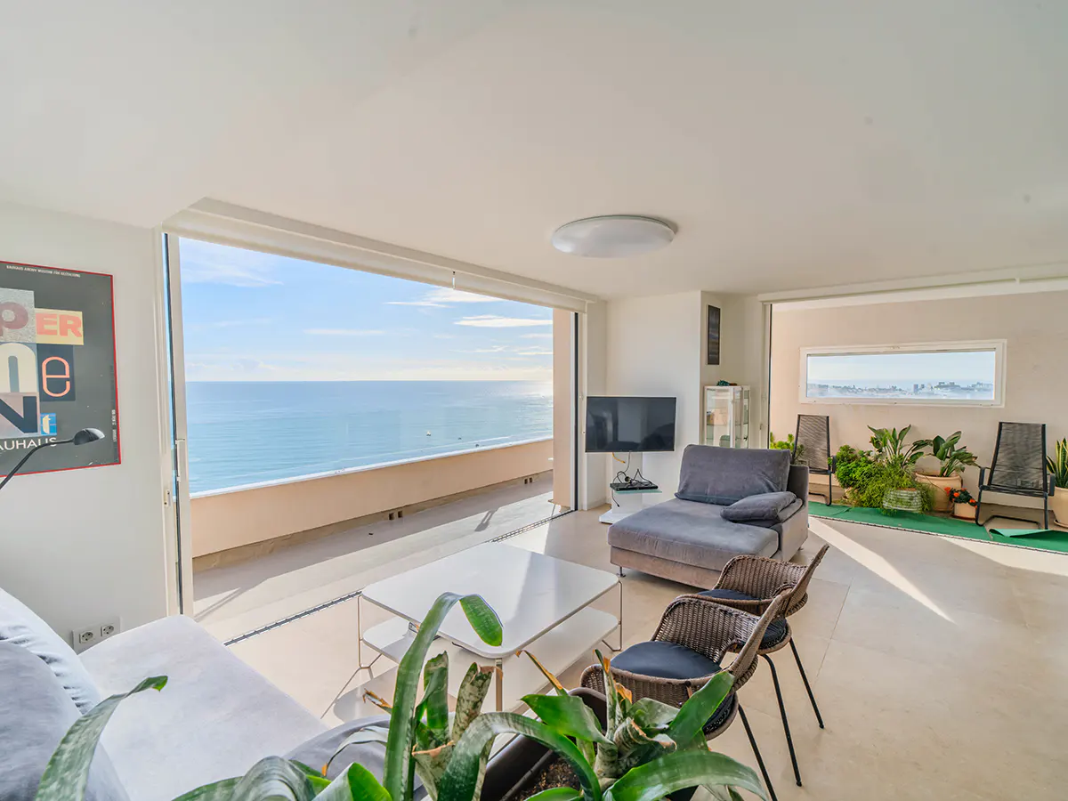 Bright living room with ocean view. Sliding glass doors open to a balcony. Gray sofa, wicker chairs, and plants furnish the space.