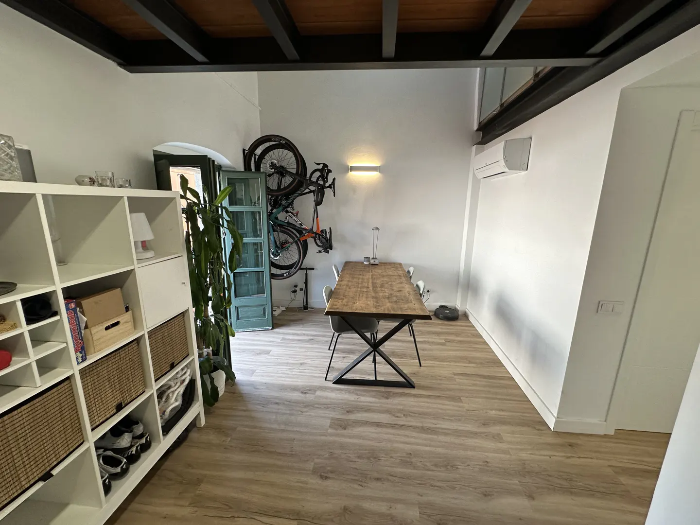Bright, modern dining room with a wood table, white walls, and a bicycle mounted on the wall. A white shelving unit is on the left.