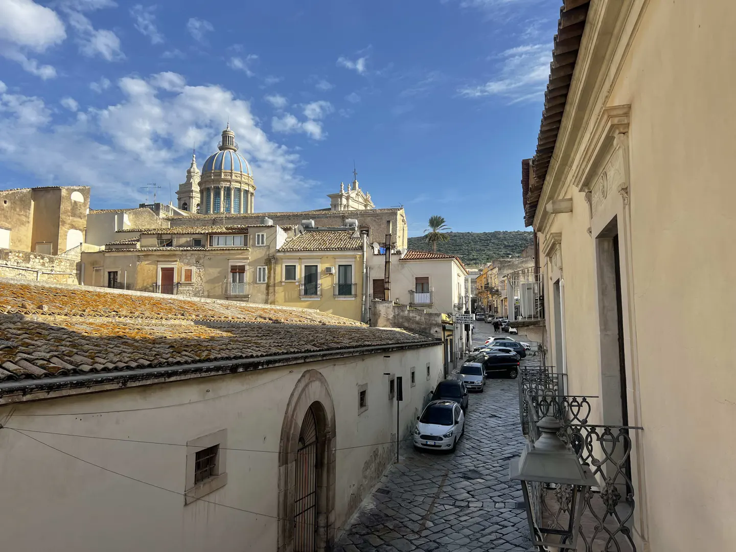 View of a European city street with cars parked along a cobblestone road, buildings, and a blue domed church in the background.
