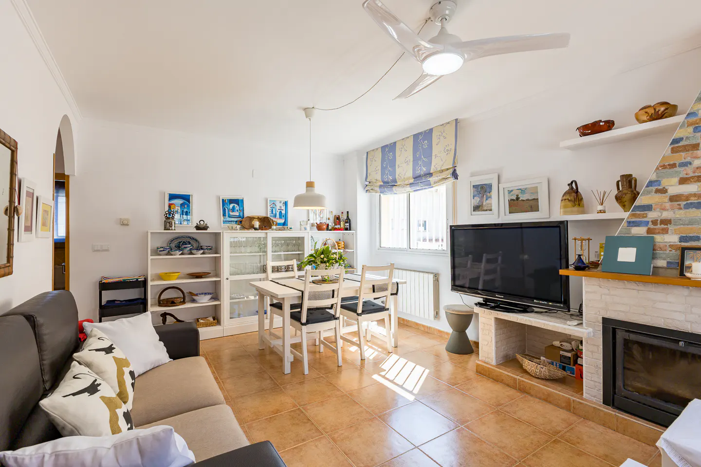 Bright living room with brown tile floor, white walls, sofa, dining table, TV, and fireplace. A ceiling fan hangs above.