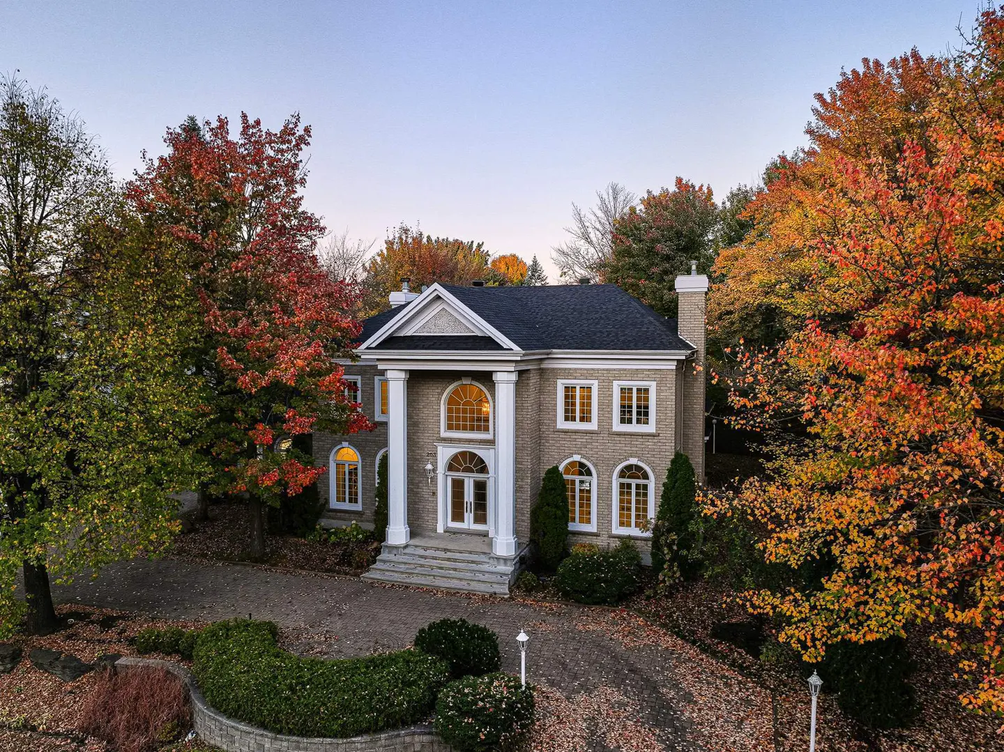 Two-story brick house with white columns and a black roof, surrounded by trees with autumn foliage.