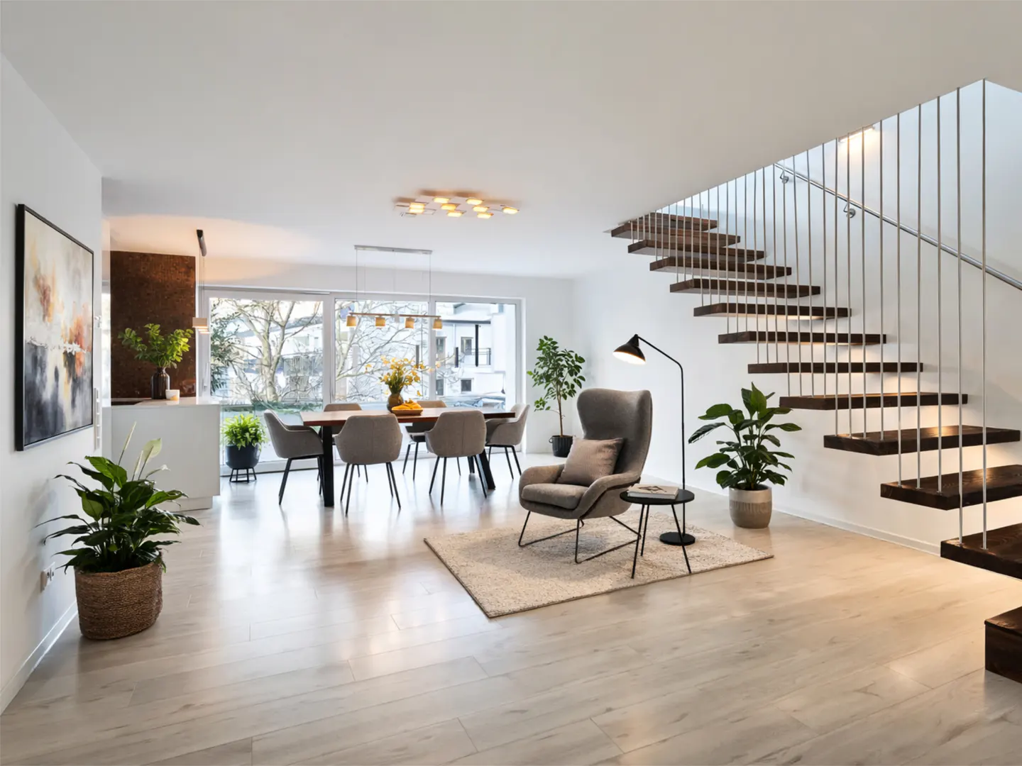 Bright, modern living room with wood floors, dining table, gray armchair, and open staircase with metal railings. Plants add a touch of nature.