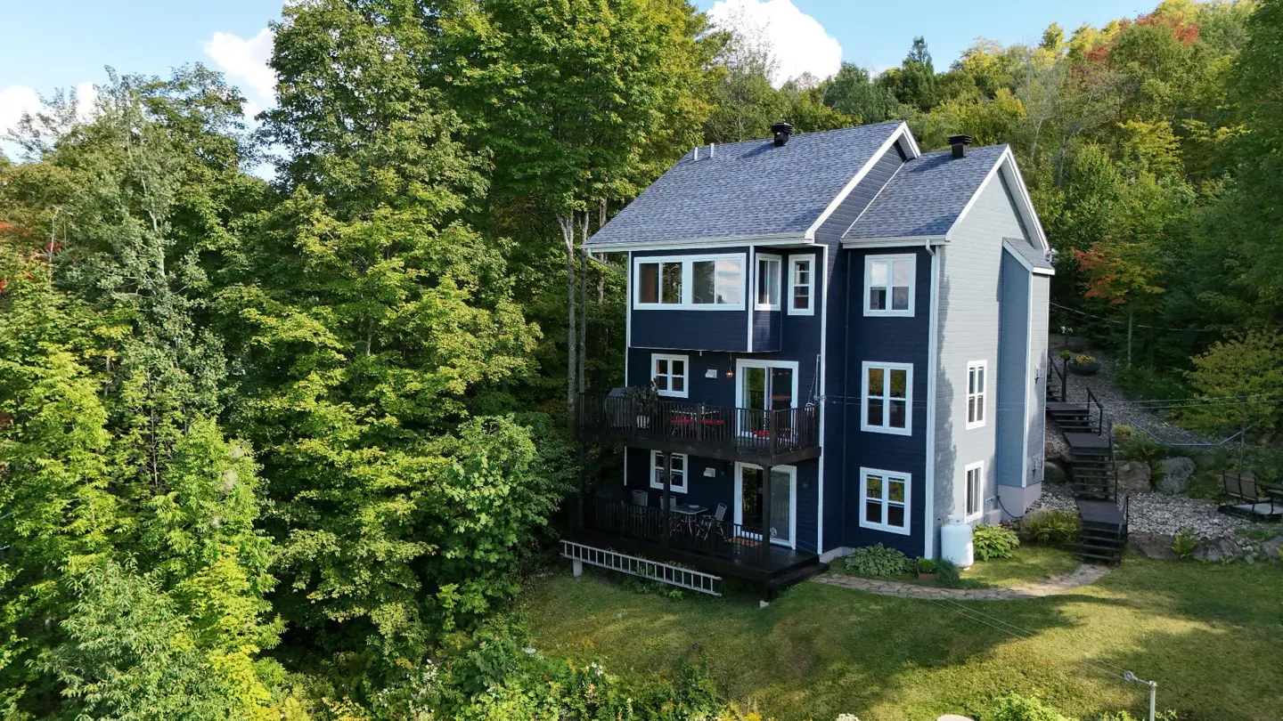 Three-story blue house with balconies, surrounded by green trees and grass.