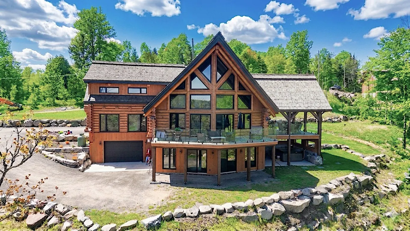 A large, modern log cabin with a wall of windows and a deck, surrounded by green trees and a blue sky.