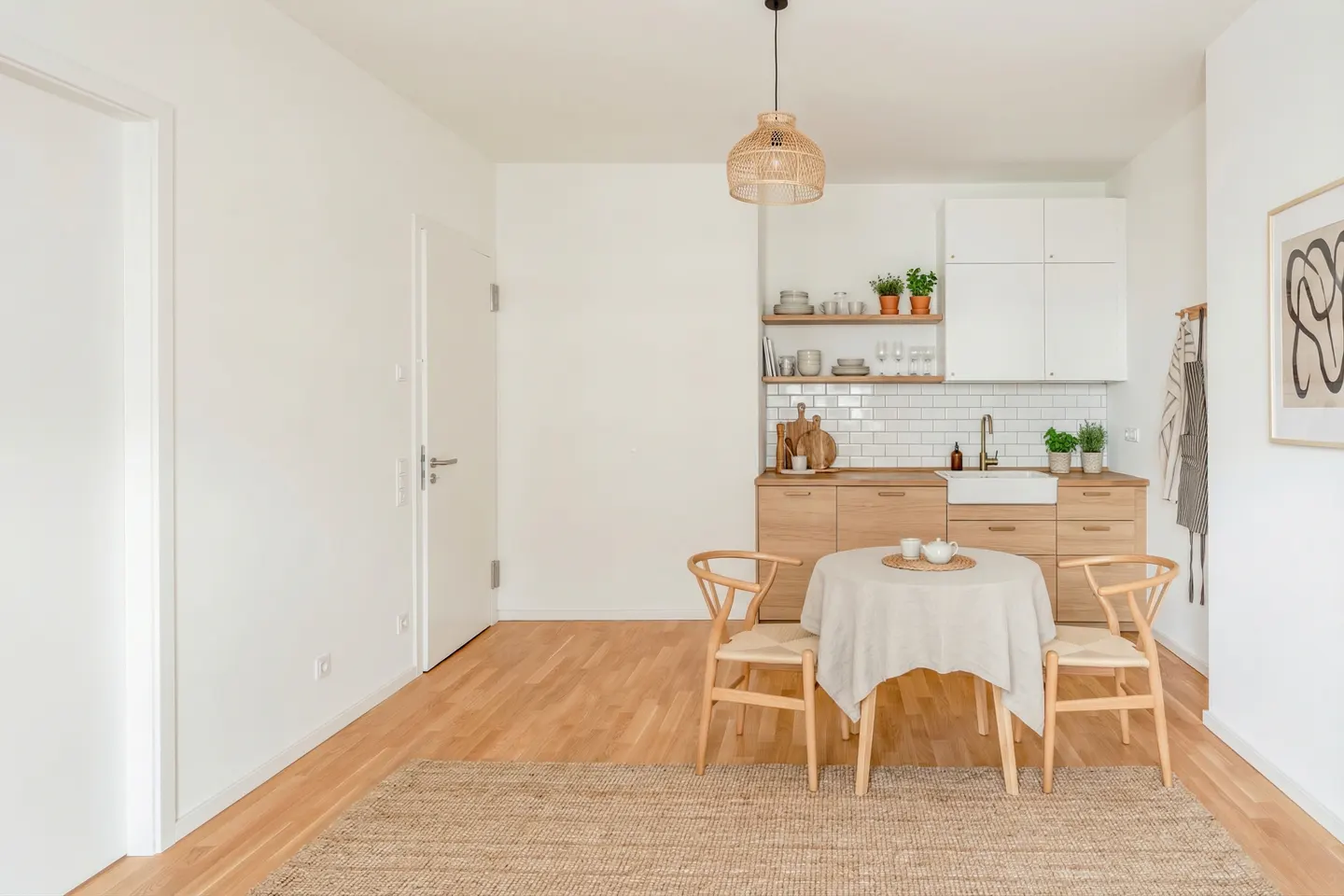 Bright, modern apartment interior with white walls, wood floors, and a jute rug. A round table with two chairs sits near a compact kitchen.