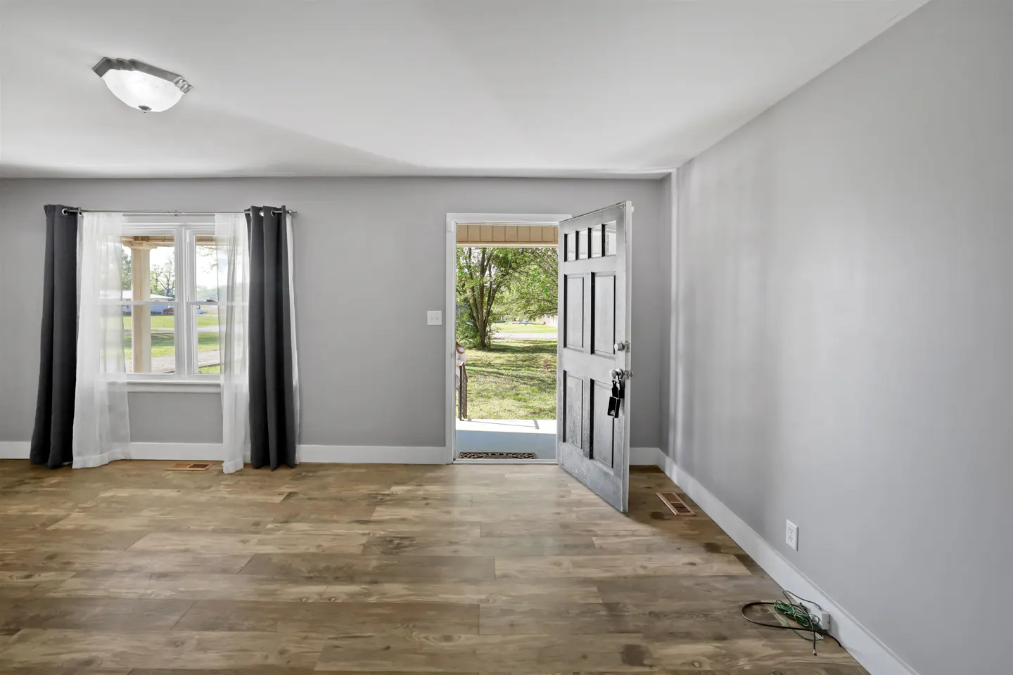 An open front door welcomes you into a gray room with wood floors and a window with gray curtains.