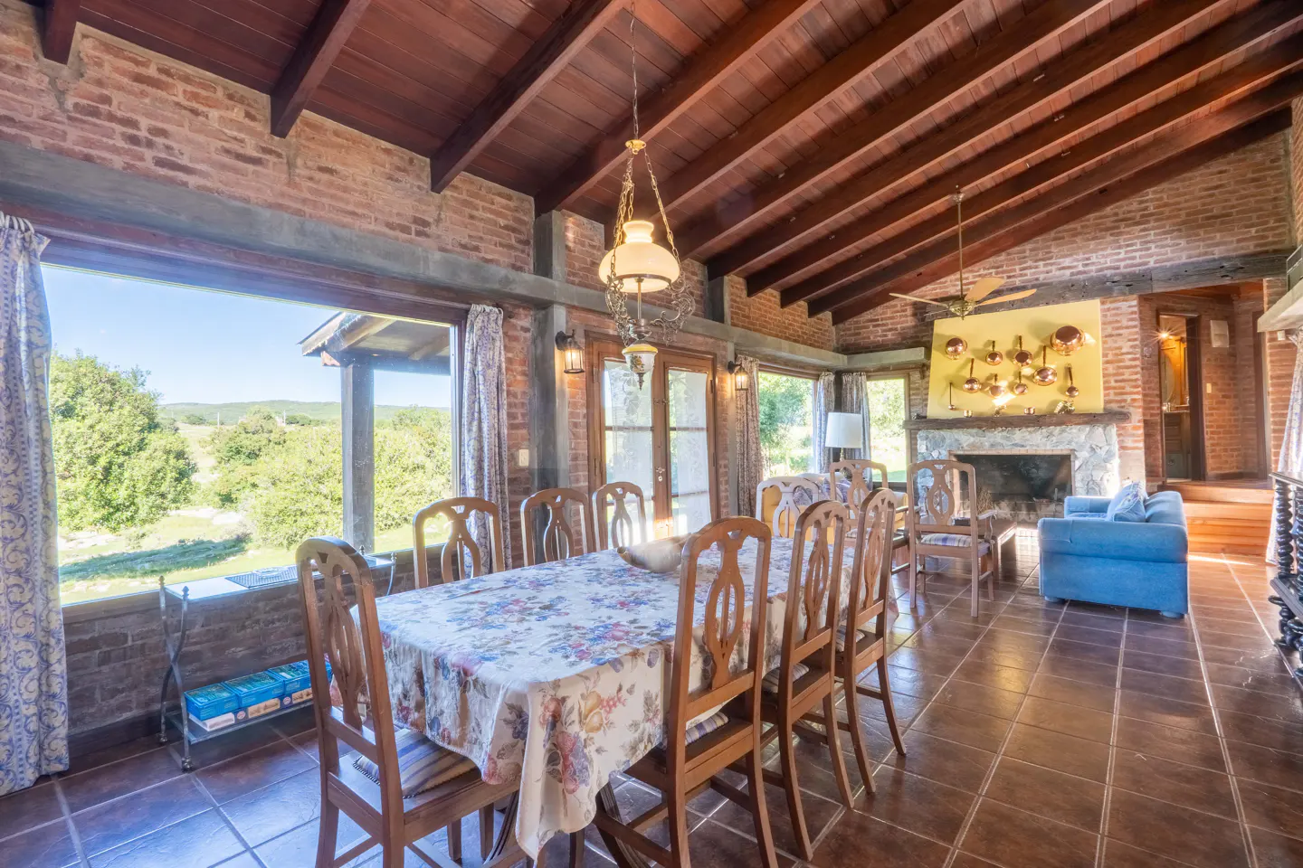 A dining room with a long table covered in a floral tablecloth, wooden chairs, brick walls, and a view of green hills.