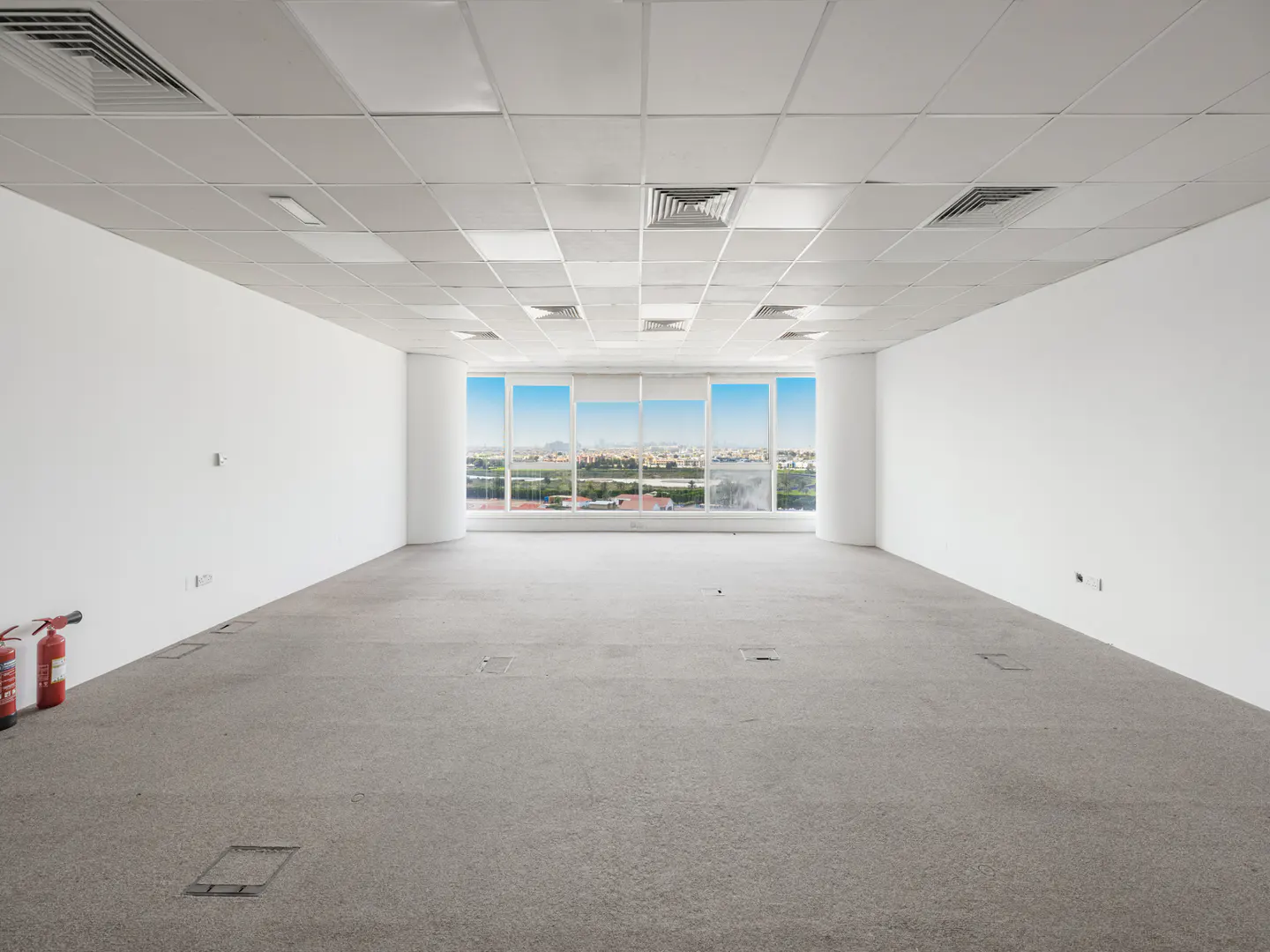 Empty office space with white walls, gray carpet, and a large window showing a city view. Two red fire extinguishers are in the corner.