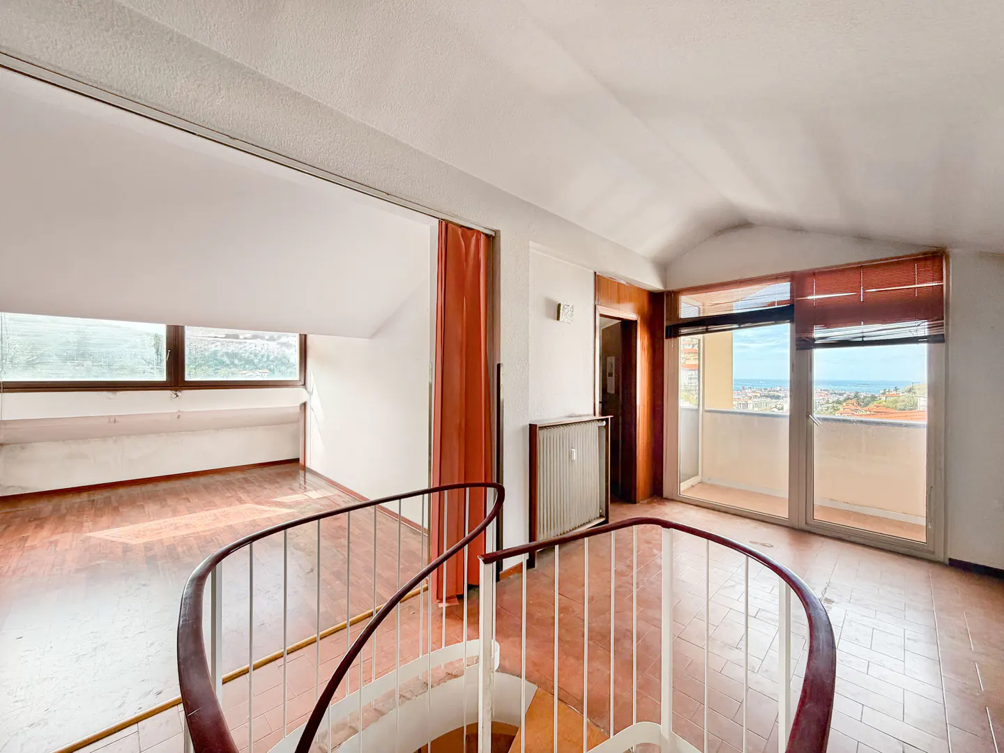 Attic room with spiral staircase, terracotta tile floor, and large windows overlooking a cityscape and ocean view.