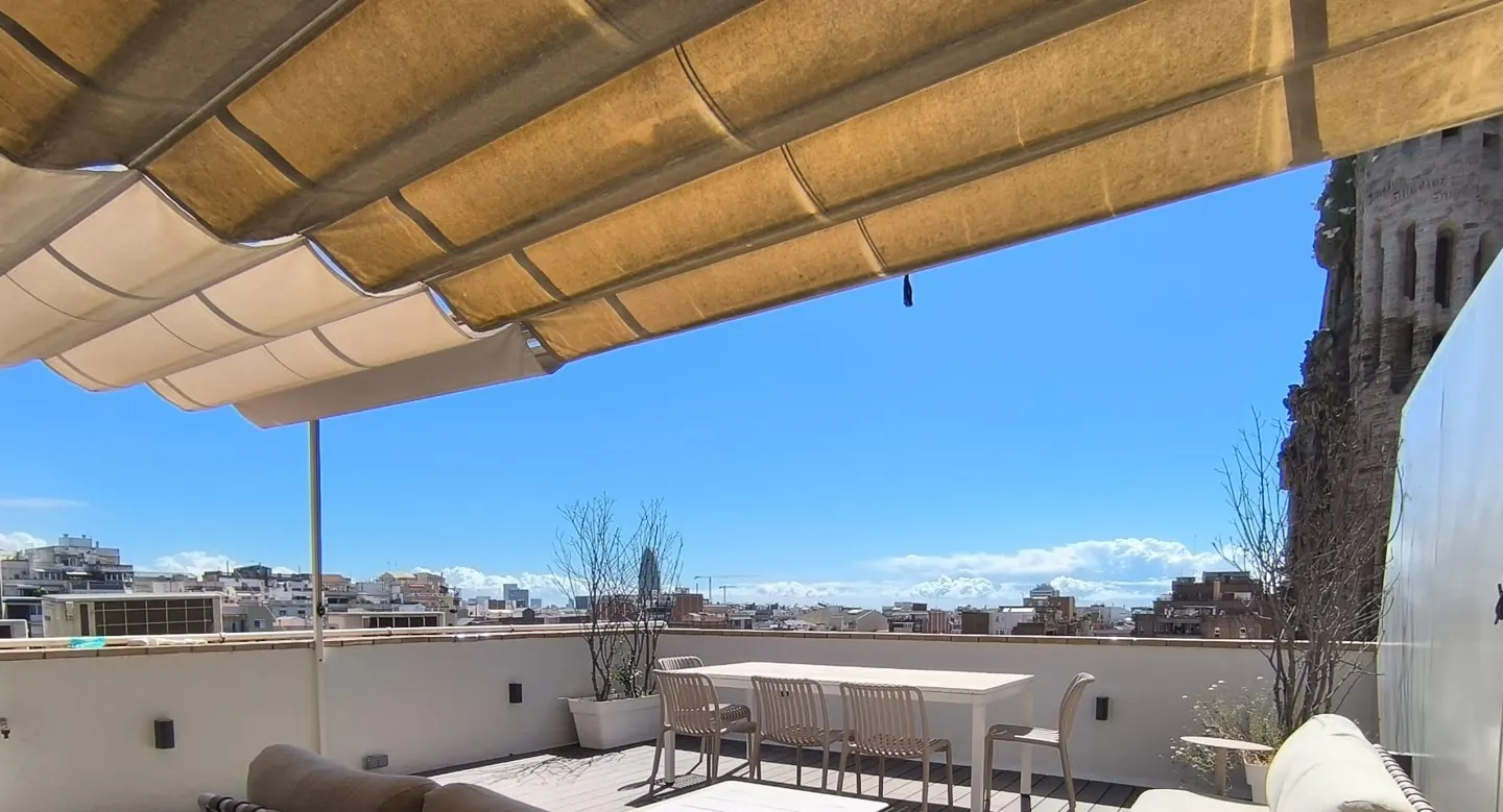 Rooftop patio with a retractable awning, white table and chairs, and city views under a bright blue sky.