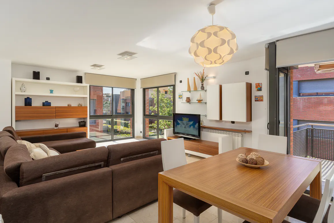 Bright living room with a brown sectional sofa, wood dining table with white chairs, and large windows overlooking a cityscape.