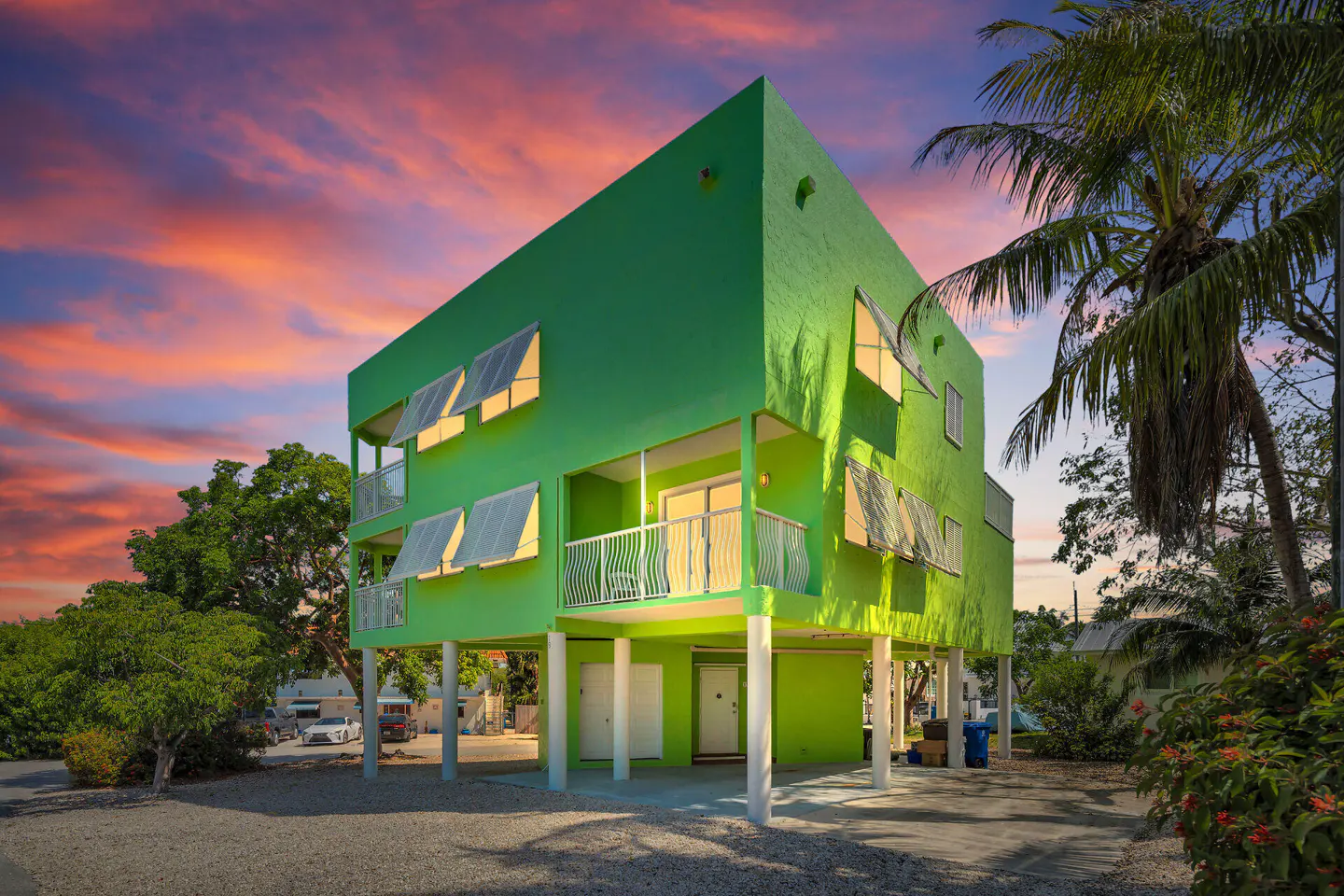 A bright green, two-story stilt house with white trim, balconies, and awnings under a colorful sunset sky.