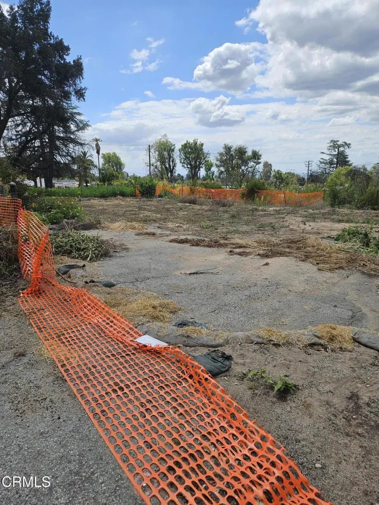 Vacant lot with orange safety fence in foreground. Trees and blue sky with clouds in background.